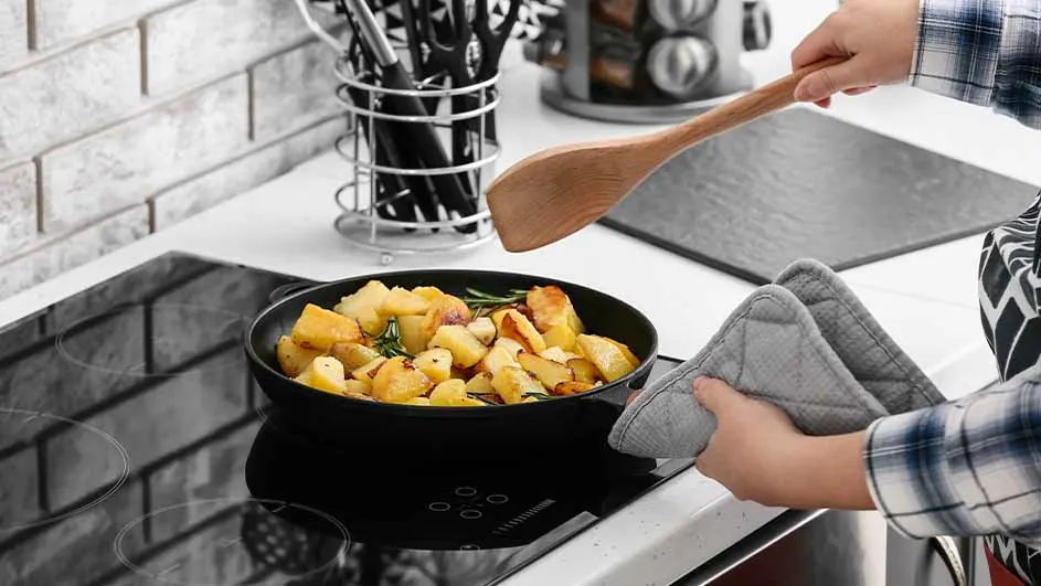 Aardappelen worden in een zwarte pan op een kookplaat gebakken, met een houten lepel en pannenlappen in de buurt.