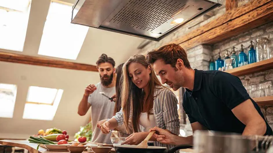 Drie mensen koken in een keuken met groenten, een kookplaat, een afzuigkap en een stenen muur met planken en blauwe flessen.