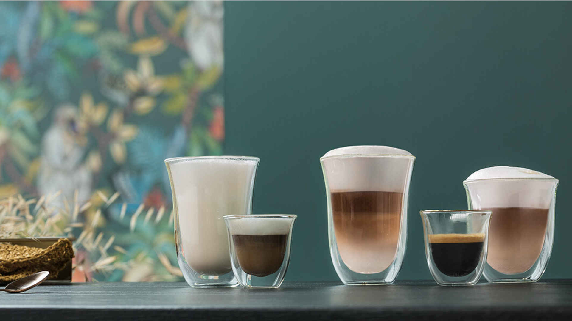 Op een tafel staan glazen met koffievariaties, zoals latte macchiato, espresso en melkkoffie. Een groenblauwe achtergrond.