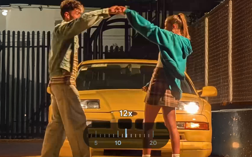 Twee personen dansen bij een gele auto 's avonds met camera zoom-interface (12x) zichtbaar. Nachtfoto met warme verlichting en stedelijke sfeer.