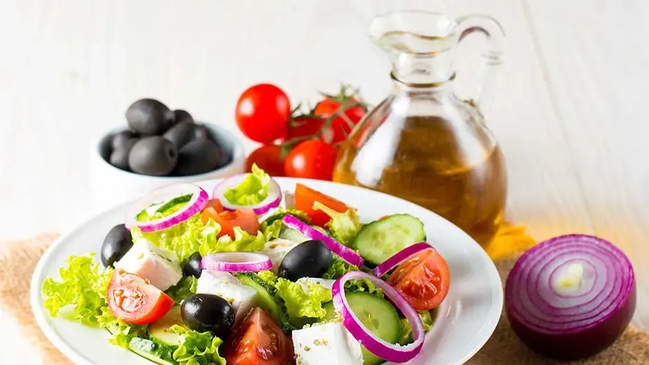 Een salade met sla, tomaat, komkommer, rode ui, feta en olijven op een wit bord, met een kan olie en extra tomaten.