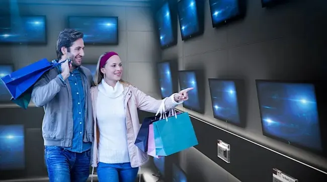 A couple with shopping bags views TVs on a store wall; she points, smiling at a screen. No brand visible.