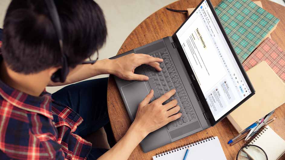 Een persoon met een headset typt op een laptop op een ronde houten tafel. Er liggen notitieblokken en pennen bij. Het scherm toont "Team Meeting".