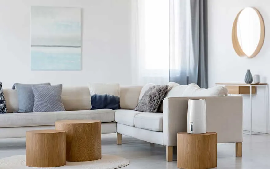 Een minimalistische woonkamer met lichtbeige bank en houten tafels, waarop een slanke witte luchtbevochtiger discreet in het interieur past.
