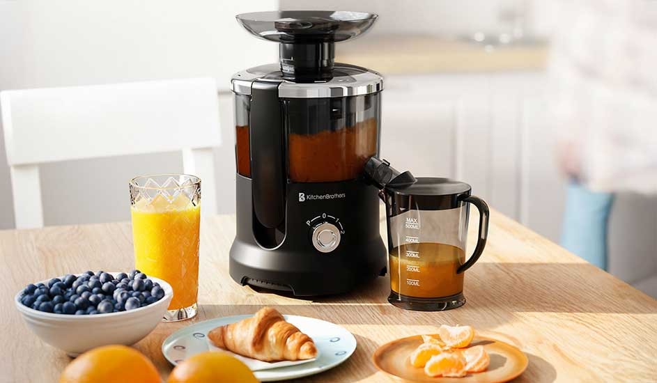A black juicer stands on a sunny wooden table with orange juice, a croissant, blueberries and peeled mandarins ready for breakfast