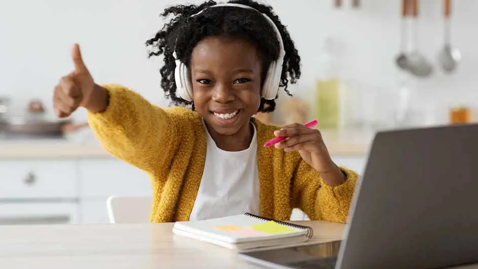 A girl in headphones at a table with a laptop, notebook and pen, gives a thumbs up in a bright room.