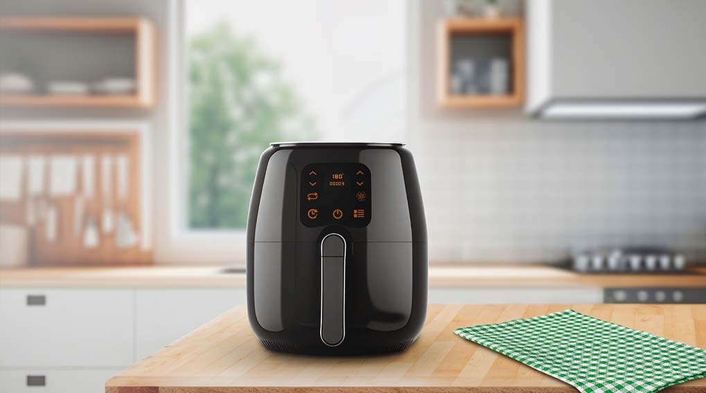 A black air fryer sits on a wooden table with a green checkered cloth. A kitchen with cabinets and a window is in the background.
