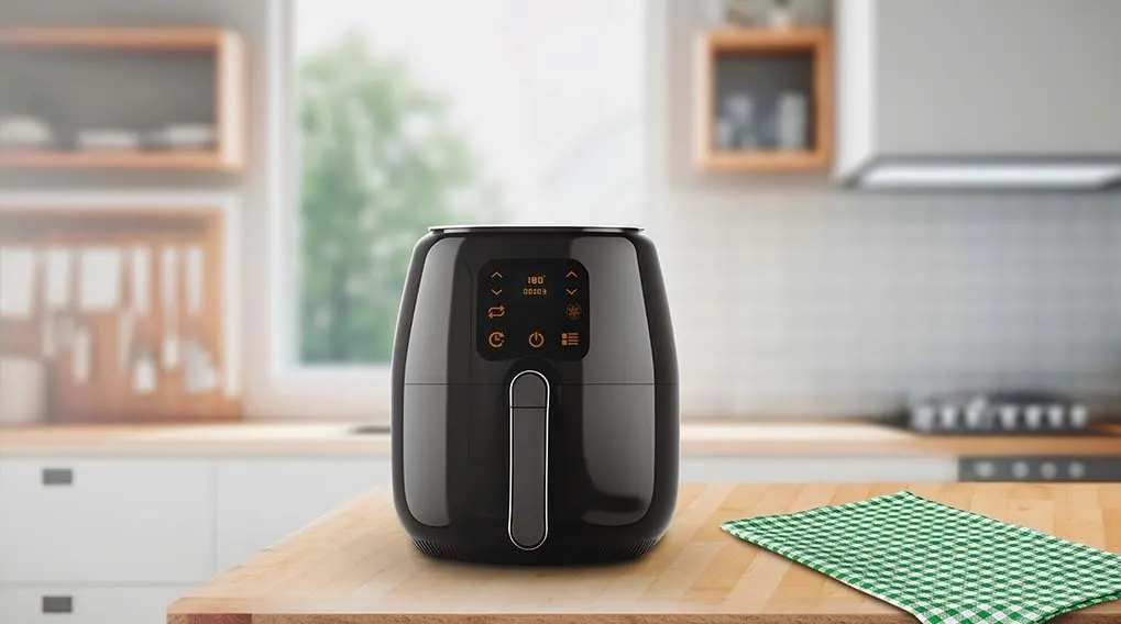 A black air fryer sits on a wooden table with a green checkered cloth. A kitchen with cabinets and a window is in the background.
