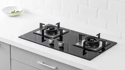 Een zwart gasfornuis met twee pitten staat op een wit aanrechtblad. Een witte kom met groene blaadjes staat linksboven.