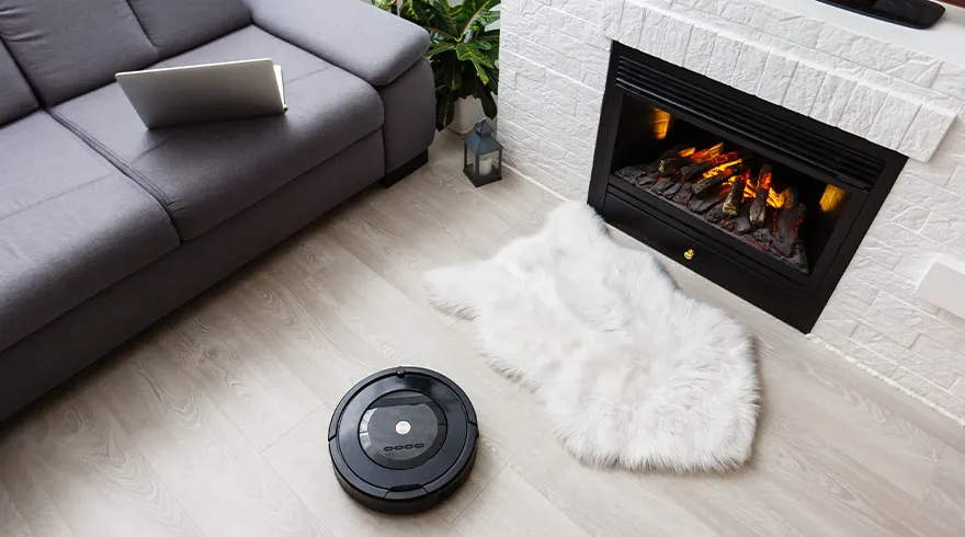 Een grijze bank met laptop, een zwarte robotstofzuiger, een wit tapijt en een witte stenen open haard.