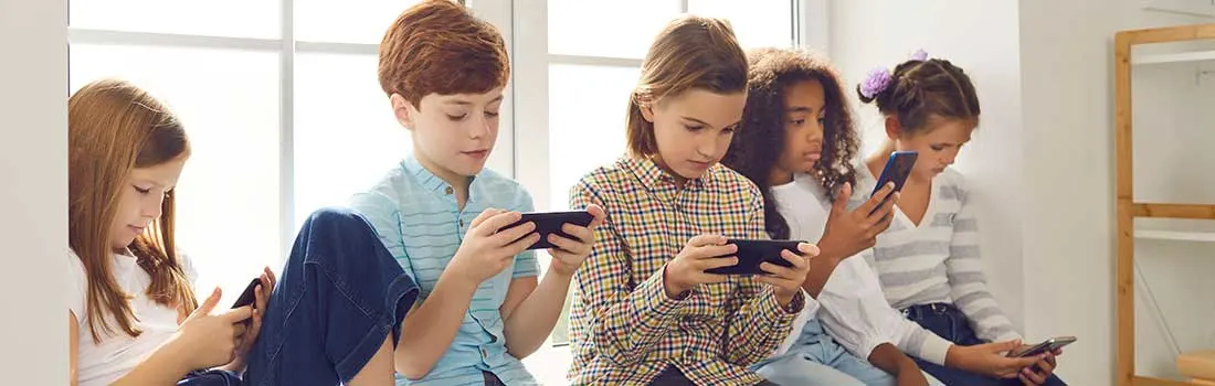 Five children sit on a windowsill, each absorbed in their own smartphone, stil en geconcentreerd op het scherm voor hen.