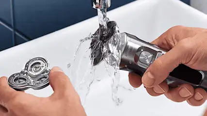 Een zilverkleurig scheerapparaat wordt onder een kraan afgespoeld, met een los scheerhoofd in de andere hand.