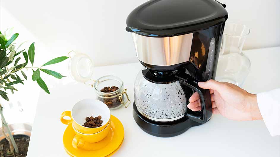 Hand holding the jug of a black filter coffee machine on a white kitchen counter, with a yellow cup, jar of coffee beans, and a small plant nearby.