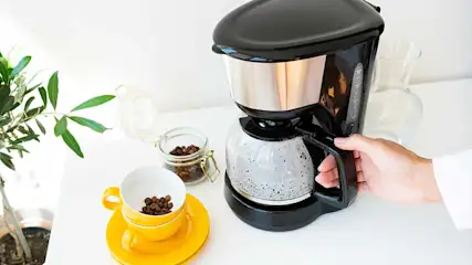 Hand holding the jug of a black filter coffee machine on a white kitchen counter, with a yellow cup, jar of coffee beans, and a small plant nearby.