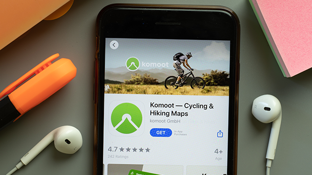 Een iPhone toont de Komoot app. Een oranje markeerstift, roze notities en witte oordopjes liggen eromheen op een grijze ondergrond.