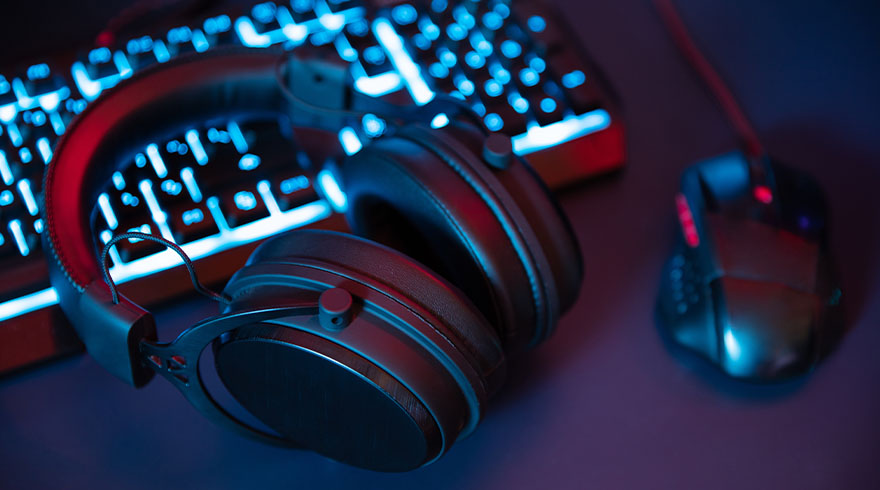 Een koptelefoon ligt voor een toetsenbord met blauwe verlichting. Een muis is rechts zichtbaar, alles op een donkere ondergrond.