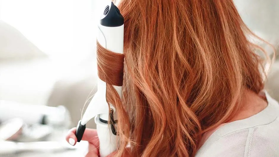 Een persoon met lang rood haar krult een lok met een witte krultang; je ziet vooral het haar en het apparaat, binnen.