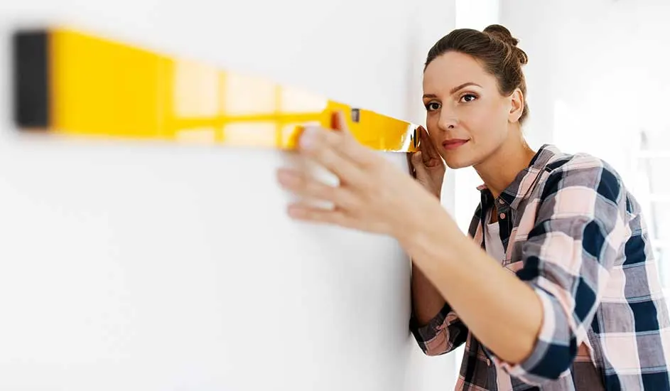 Iemand in een geruite blouse houdt een gele waterpas tegen een witte muur om te controleren of deze recht hangt in een lichte kamer.