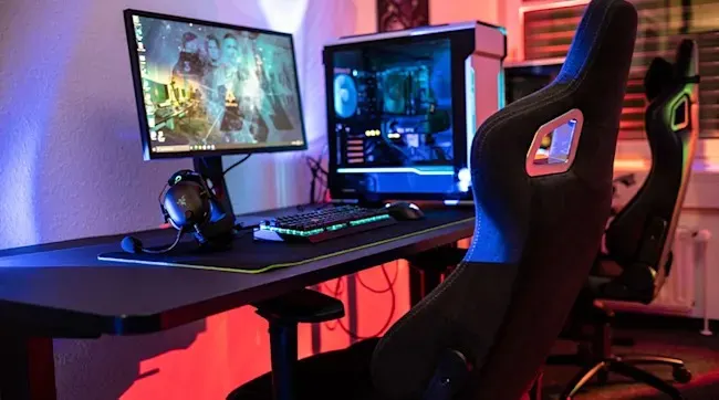 Dark gaming setup with monitor, glowing PC case, RGB keyboard and mouse, surrounded by two ergonomic gaming chairs.