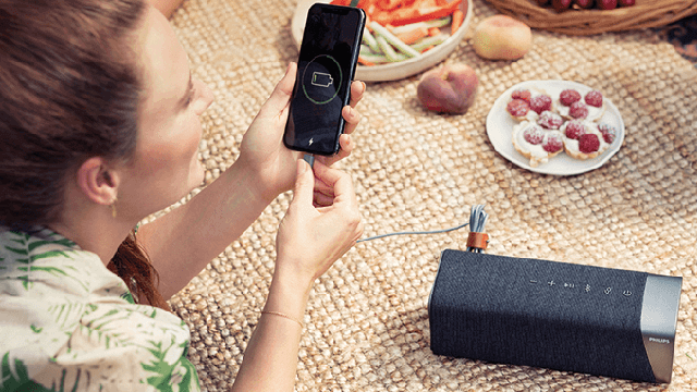 Vrouw verbindt smartphone met grijze speaker op een kleed, met eten in de achtergrond. Telefoon toont batterij-icoon.