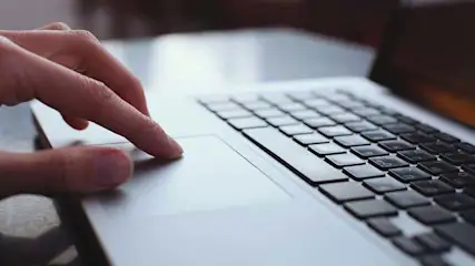 Een hand tikt op een touchpad van een zilverkleurige laptop, met zwarte toetsen zichtbaar op het toetsenbord.
