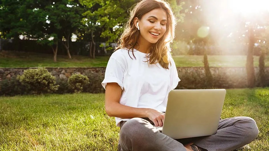 A smiling woman sits on grass in a park with a laptop and earbuds; warm sunlight and trees behind her.