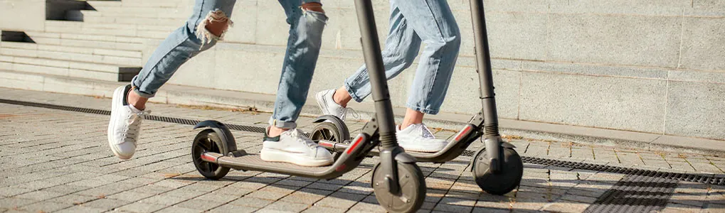 Two people riding scooters on a paved surface, wearing jeans and sneakers, with steps visible in the background.