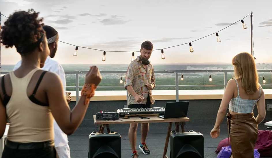 Rooftop party with a DJ setup on a wooden table, two speakers, and string lights. Guests dancing with a scenic view of the green city beyond.