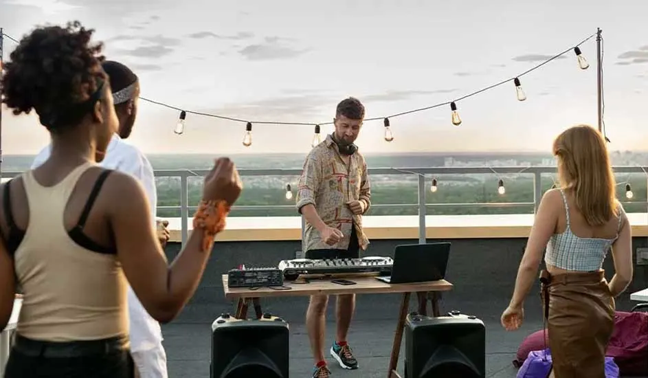 Rooftop party with a DJ setup on a wooden table, two speakers, and string lights. Guests dancing with a scenic view of the green city beyond.