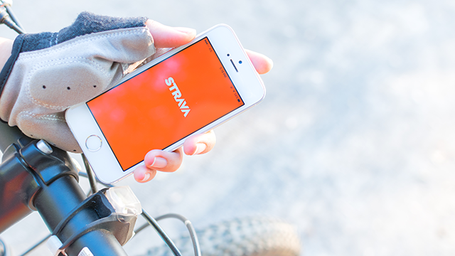 Een hand met een fietshandschoen houdt een witte smartphone vast met de Strava-app op een fietsstuur.