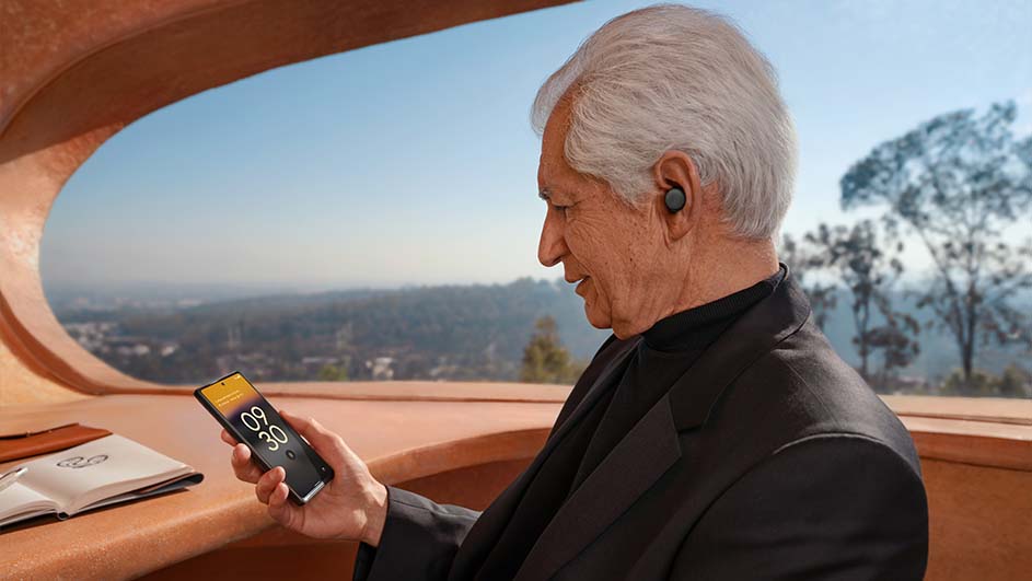 Oudere man met oortjes kijkt naar een telefoon met de tijd 09:30. Op de achtergrond is een landschap zichtbaar door een raam.