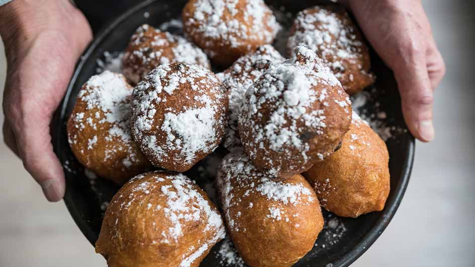 Twee handen houden een zwarte schaal vol gefrituurde oliebollen, bedekt met poedersuiker.