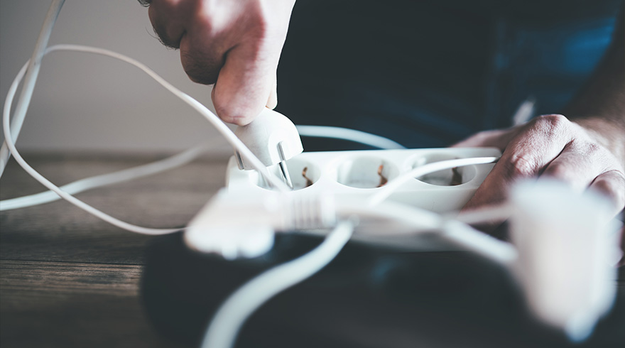 Een hand steekt een witte stekker in een wit stekkerblok met meerdere stopcontacten, op een donkere ondergrond.