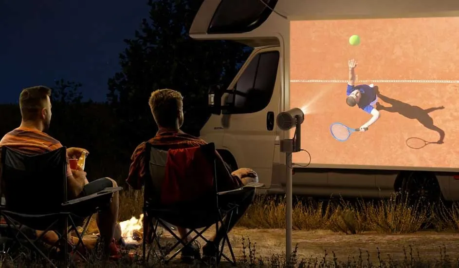 Two people sit outdoors in front of a camper van, watching a tennis match projected onto its side by a Samsung portable projector at night.
