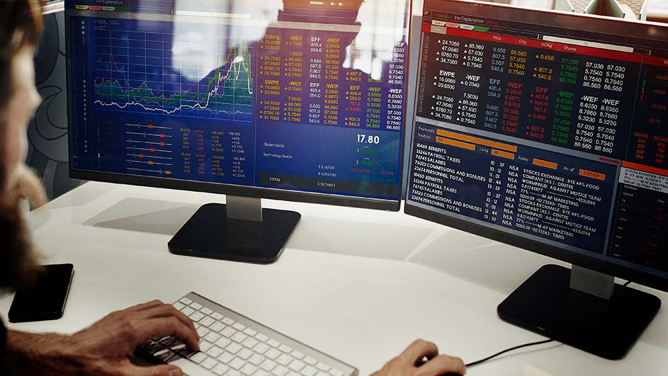 Twee computerschermen tonen financiële data en grafieken. Handen typen op een toetsenbord, met een witte tafel en een zwarte telefoon.