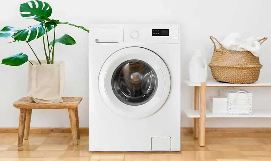 Een witte wasmachine staat in een lichte kamer naast een plant en mand; de ruimte oogt fris, rustig en verzorgd ingericht.