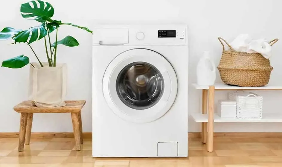Een witte wasmachine staat in een lichte kamer naast een plant en mand; de ruimte oogt fris, rustig en verzorgd ingericht.