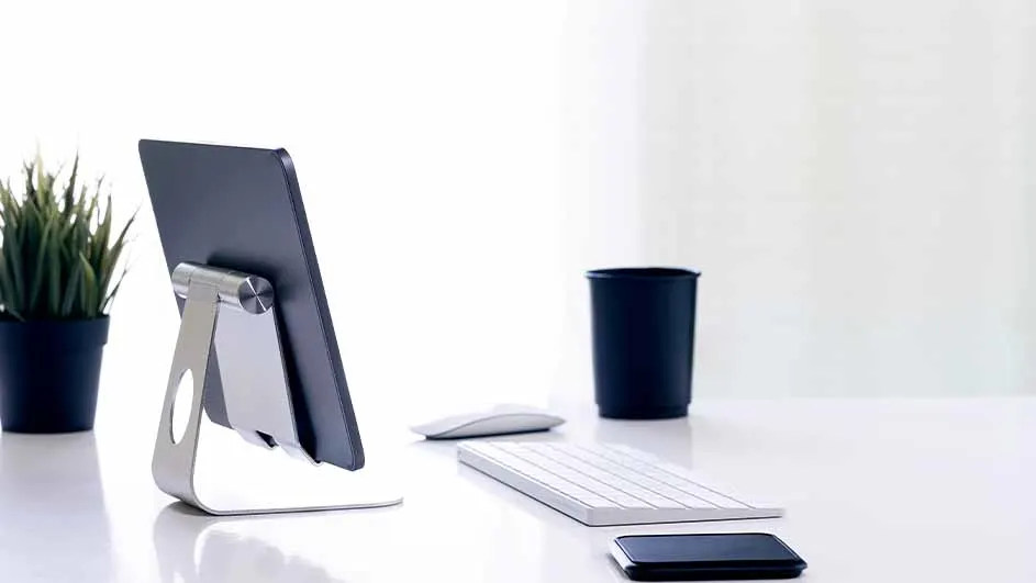 Op een wit bureau staan een tablet op een zilveren standaard, een toetsenbord, een muis, een zwarte beker en een plant.