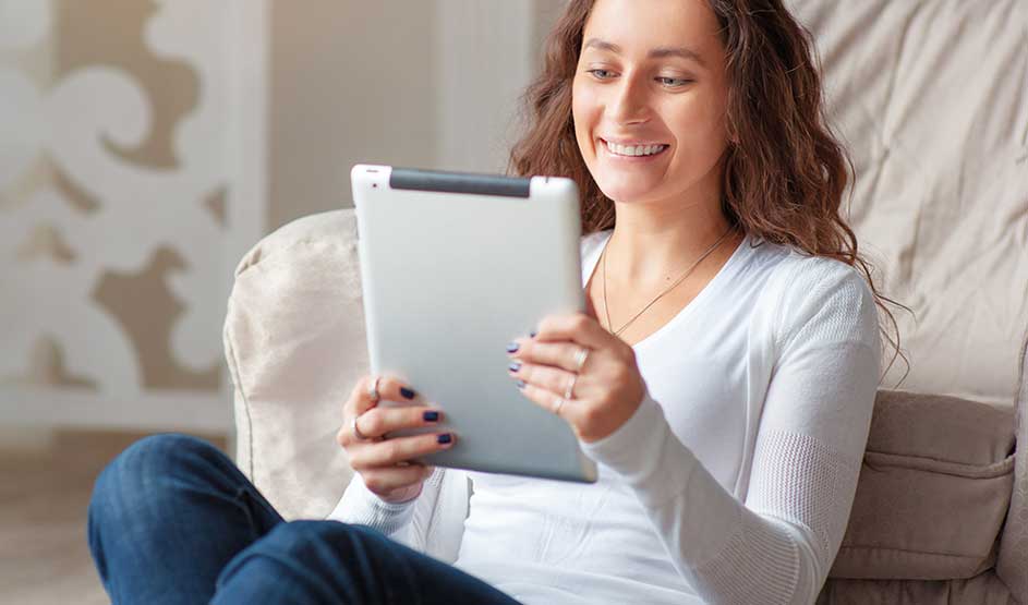 Een vrouw zit ontspannen op de bank en glimlacht terwijl ze een tablet in haar handen houdt, verdiept in lezen of een gezellige videogesprek.