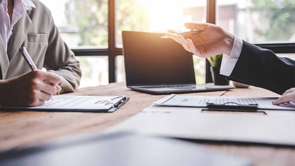 Twee personen aan een tafel met documenten en een laptop. Een persoon schrijft, de ander wijst met een pen.