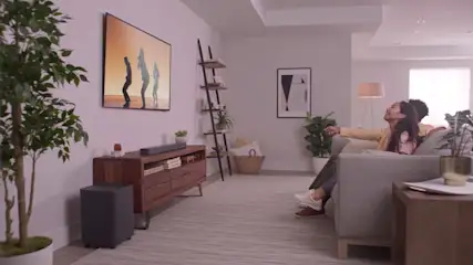 Een woonkamer met een tv, een paar mensen op een grijze bank en een houten kast.