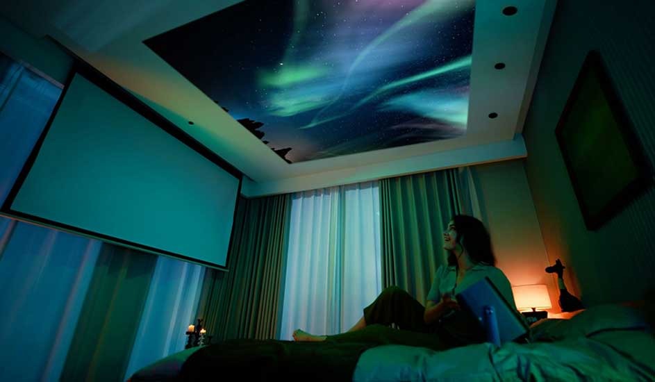 A woman sits on her bed in a softly lit room, watching a vivid aurora borealis projected onto the ceiling. No brand name is visible in the scene.