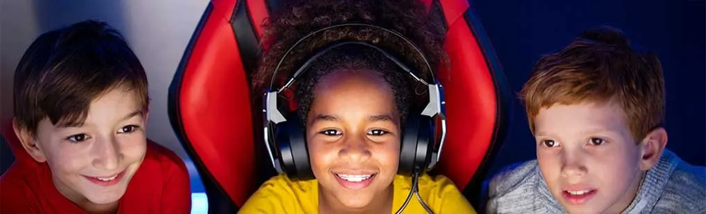 Three children sit together in front of a screen, one wearing a headset and sitting in a red game chair.