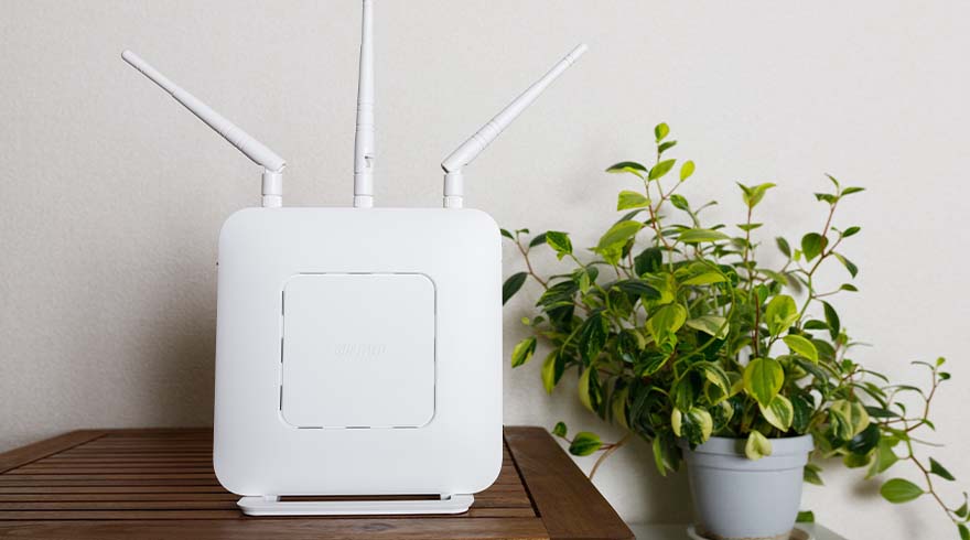 Een witte router met drie antennes staat op een houten tafel, naast een potplant met groene bladeren.