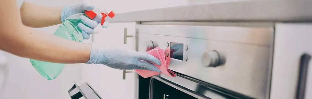 Hands in blue gloves clean a stainless steel oven with a pink cloth and a green spray bottle.