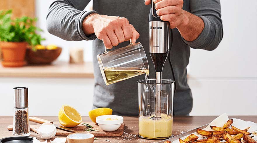 Een persoon gebruikt een staafmixer in een beker, terwijl ze olie uit een glazen kan gieten. Er liggen ingrediënten en frietjes.