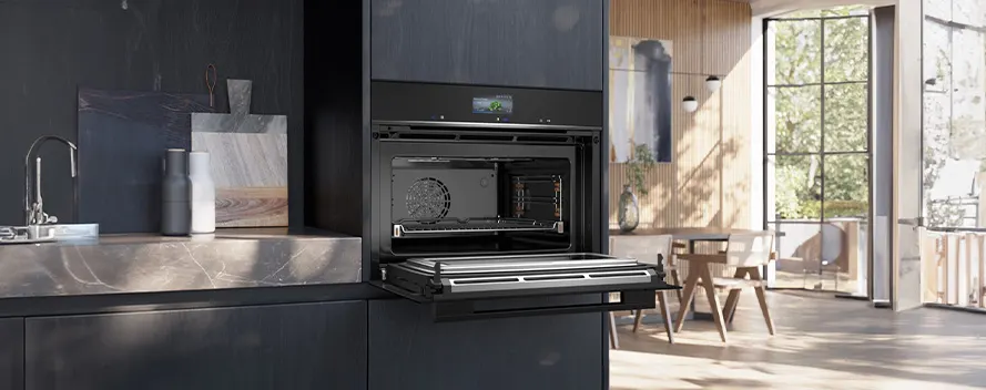 A kitchen features an open oven on dark cabinetry, a metal countertop with a sink, and a sunlit dining area.