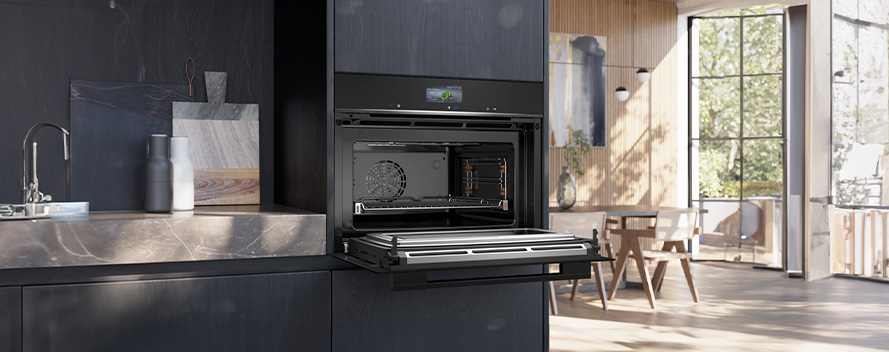 Moderne keuken met zwarte kasten, een openstaande oven en een aanrecht met kraan en snijplanken. Eethoek met ramen op achtergrond.