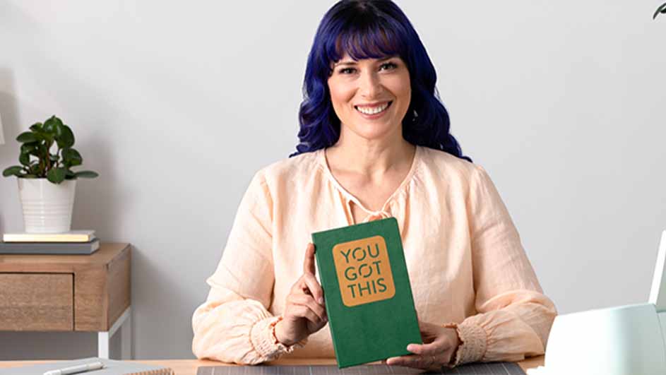 Vrouw houdt groen boek met de tekst "You Got This" in gouden letters vast, zittend aan een tafel met een plant op de achtergrond.