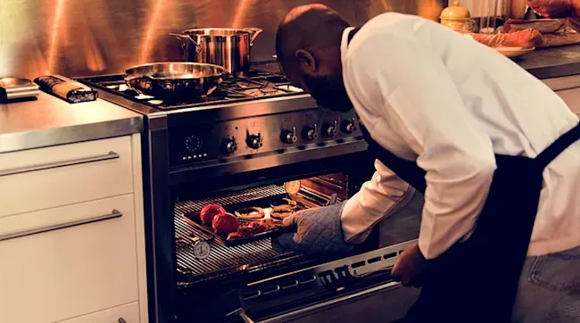 A chef wearing a white shirt and a black apron is opening a silver oven with food inside, in a commercial kitchen.